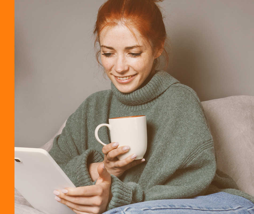 Eine Frau sitzt mit einer Tasse in der Hand auf dem Sofa und schaut auf ihr Tablet.