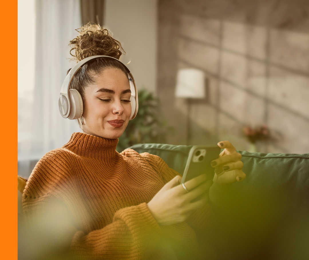 Eine Frau sitzt mit Kopfhörern auf einem Sofa und hat ihr Handy in der Hand.
