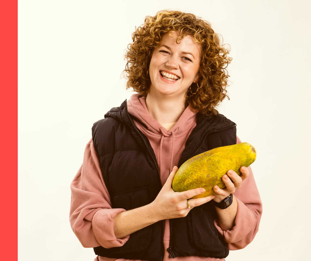 Eine Frau hält eine Papaya in der Hand und lacht in die Kamera.
