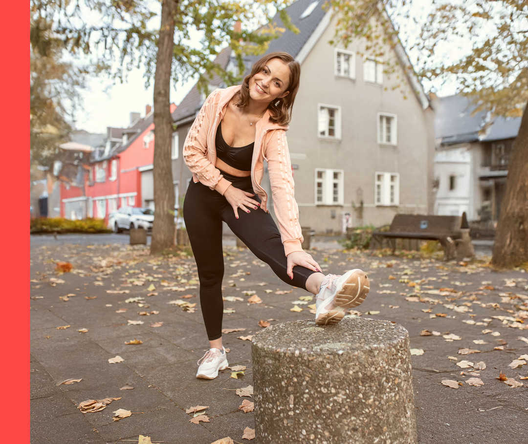 Anna dehnt in Sportkleidung ihr Bein und lächelt im Freien in die Kamera.