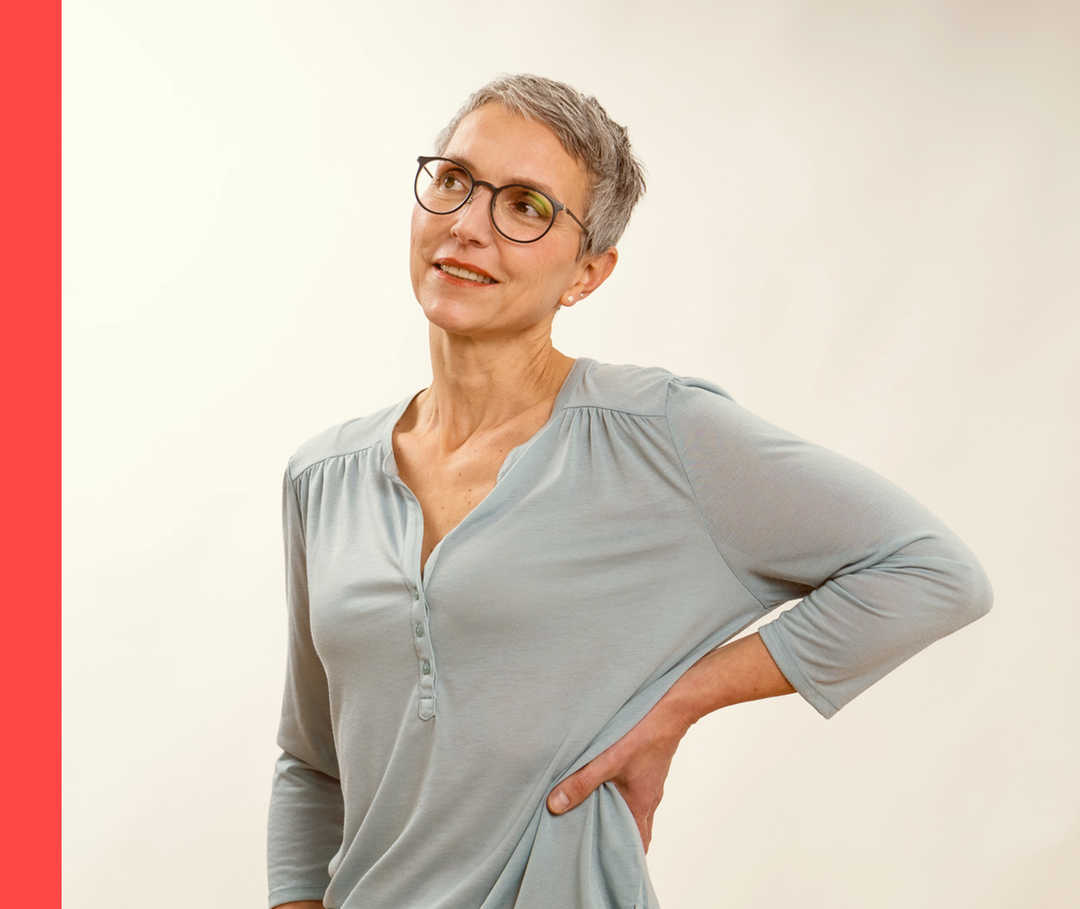 Eine Frau in einer locker sitzenden Bluse stemmt die linke Hand in die Hüfte.