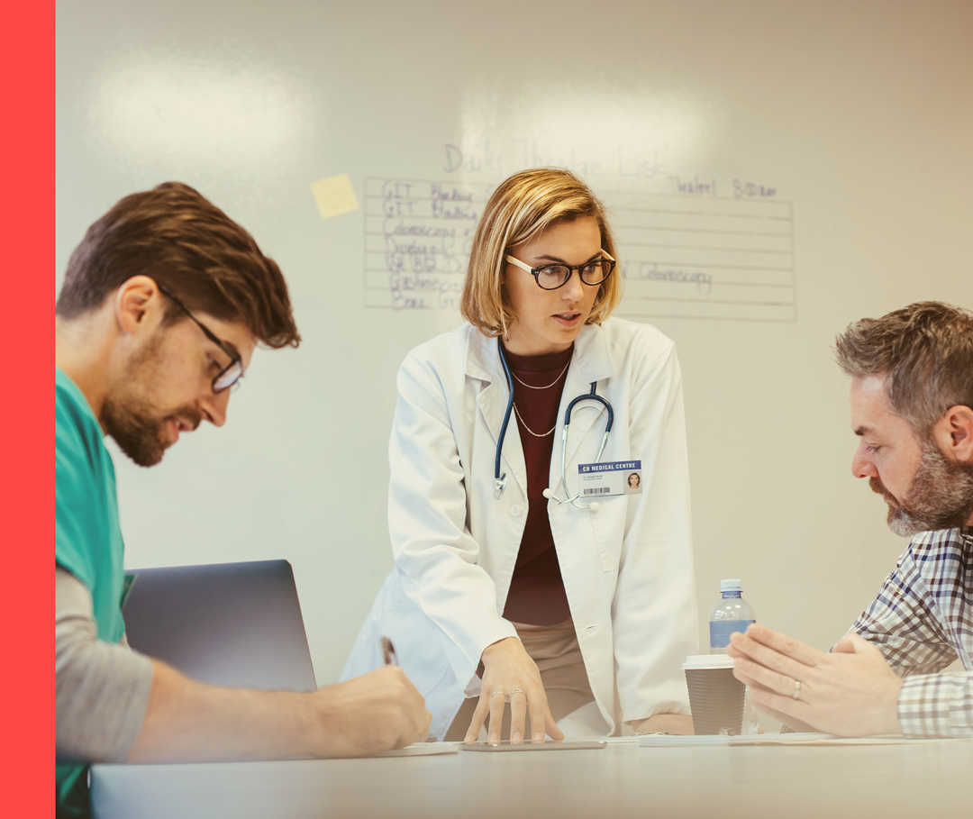 Eine Frau in weißem Kittel steht an einem Tisch und unterhält sich mit mehreren sitzenden Personen.