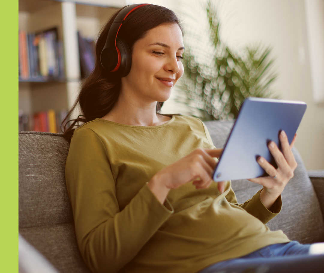 Eine Frau mit Kopfhörern sitzt auf einer Couch und hält ein Tablet in der Hand.