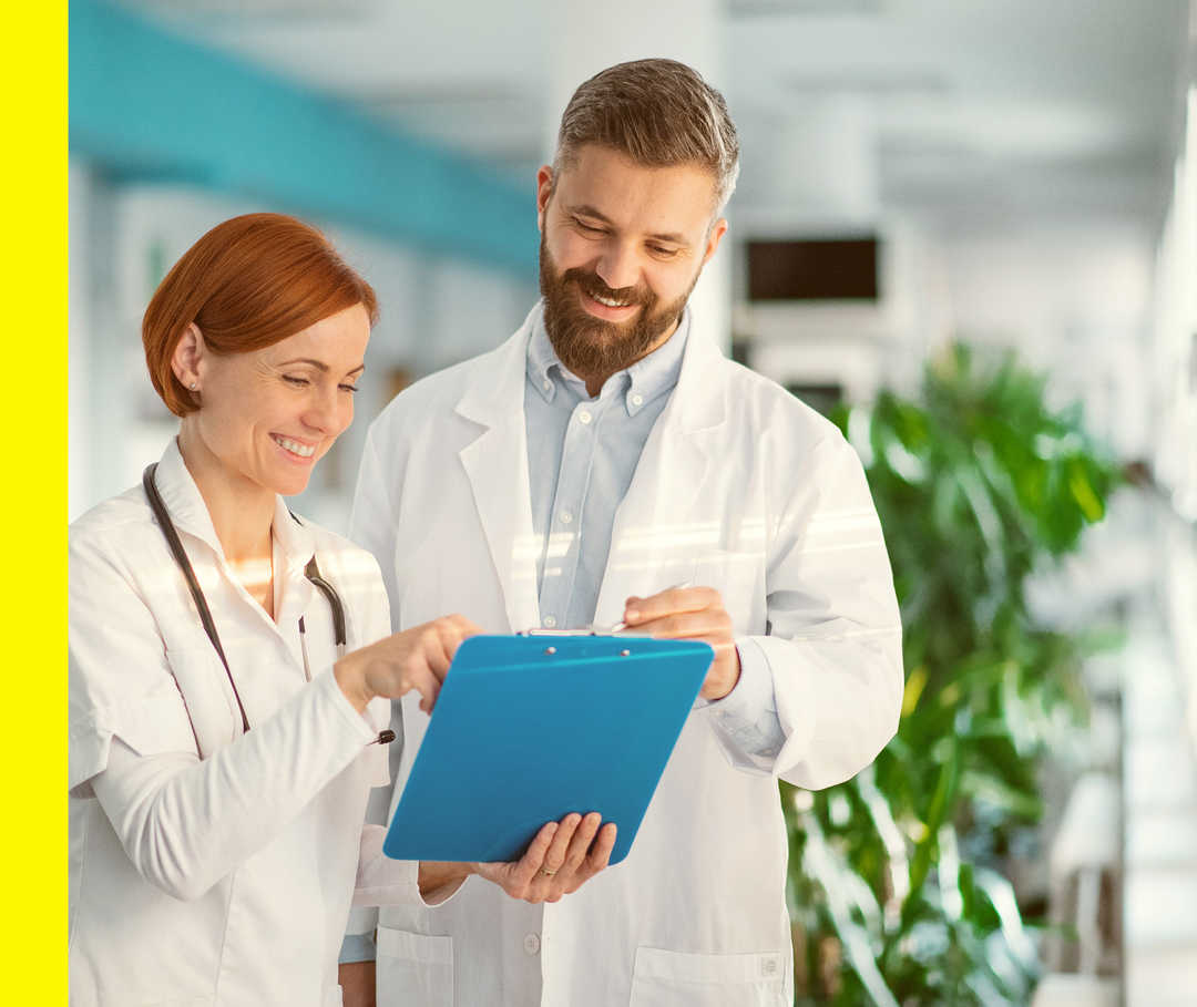 Eine Frau und ein Mann im weißen Kittel schauen lächelnd auf ein Klemmbrett.