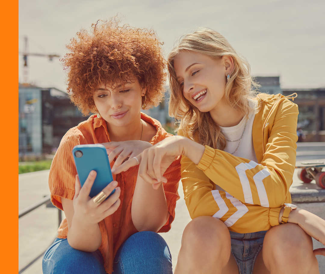 Zwei junge Personen sitzen nebeneinander und schauen lächelnd auf ein Smartphone.