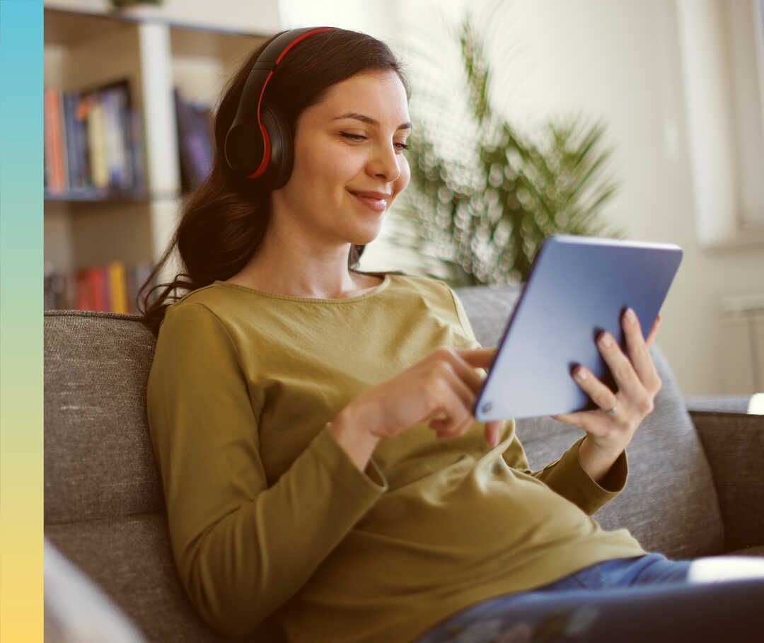 Eine Frau mit Kopfhörern sitzt auf einer Couch und hält ein Tablet in der Hand.